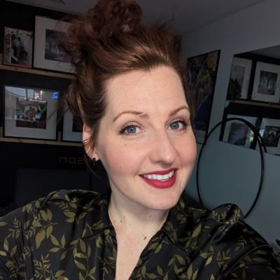 一位女性微笑，头发蓬松，穿着花卉图案的上衣，背景中有艺术作品和镜子。